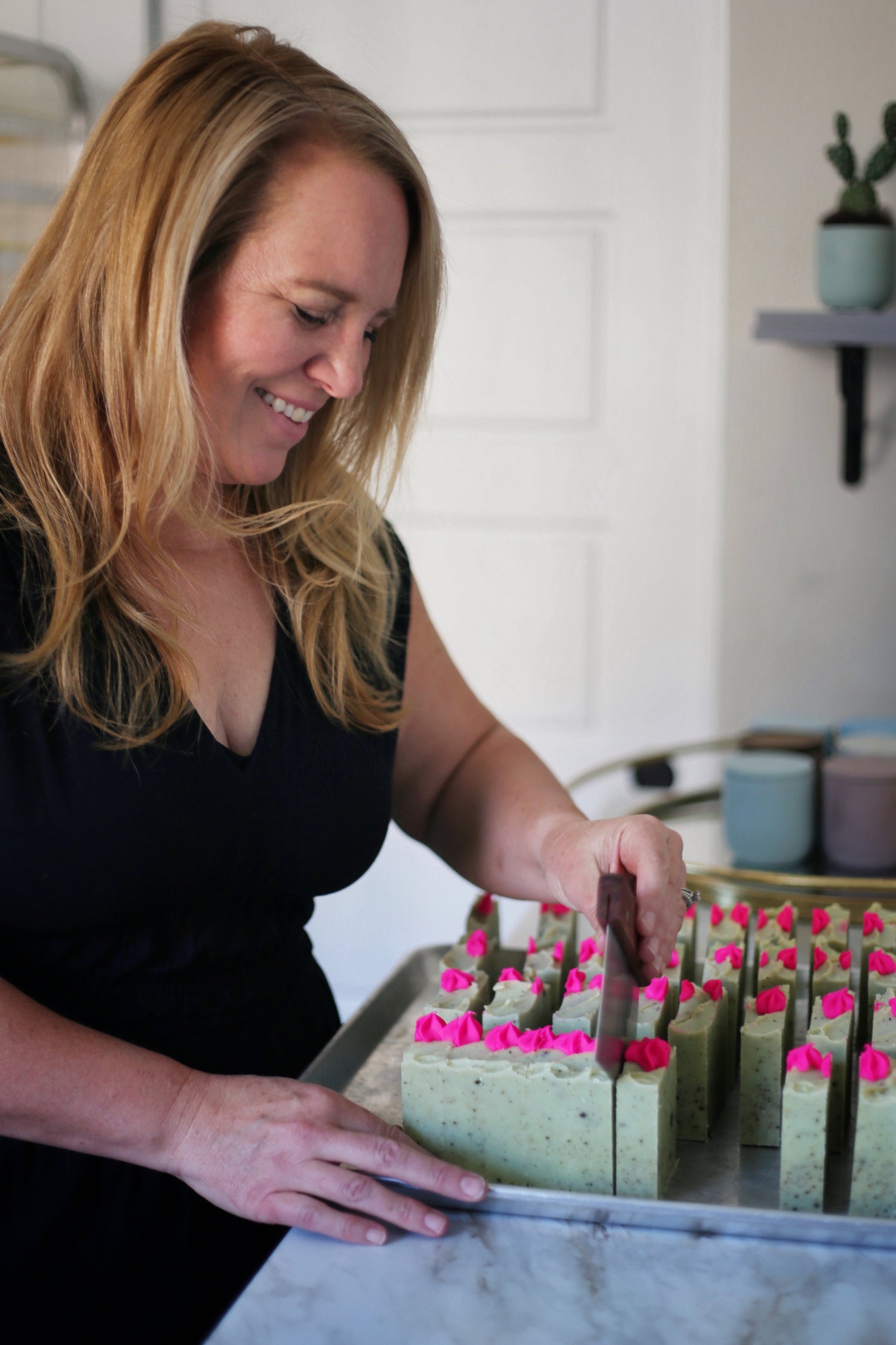 Woman hand-cutting freshly made green artisan soap topped with bright pink frosting-style swirls on a tray in a home studio.